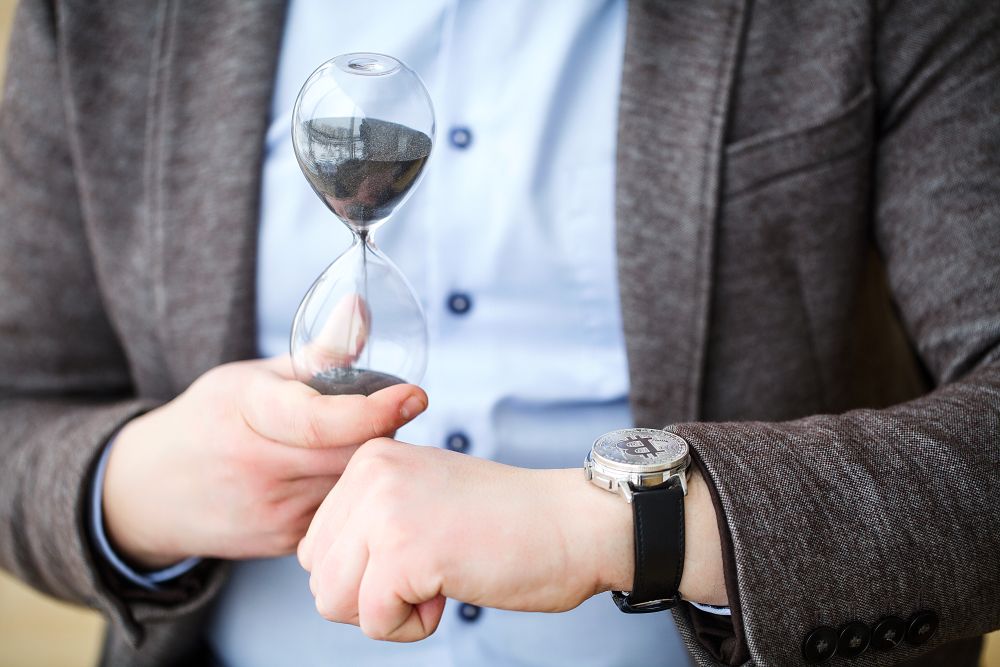 Mann zeigt Armbanduhr und Sanduhr in Richtung der Kamera